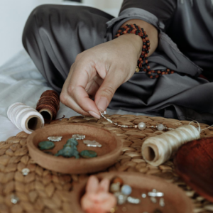 A person sits indoors engaged in crafting jewelry using beads and thread.