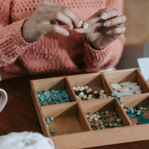 Crop unrecognizable ethnic female in light orange sweater doing beading with colorful beads and fishing line